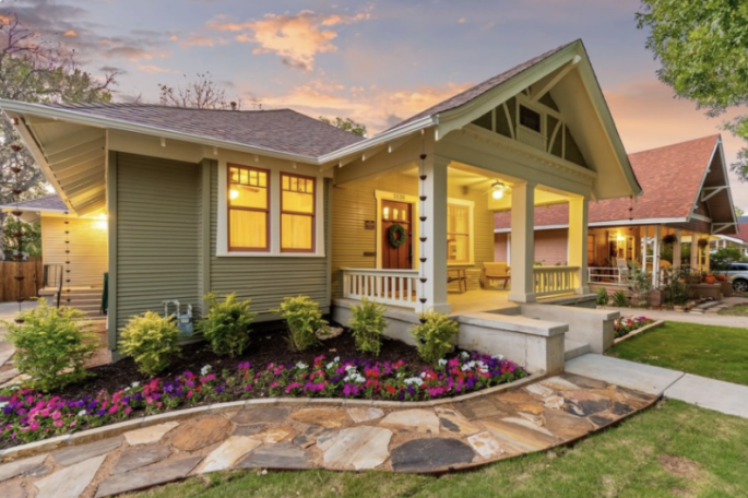 Charming green house with a porch, landscaped gardens, and colorful flowers at sunset.