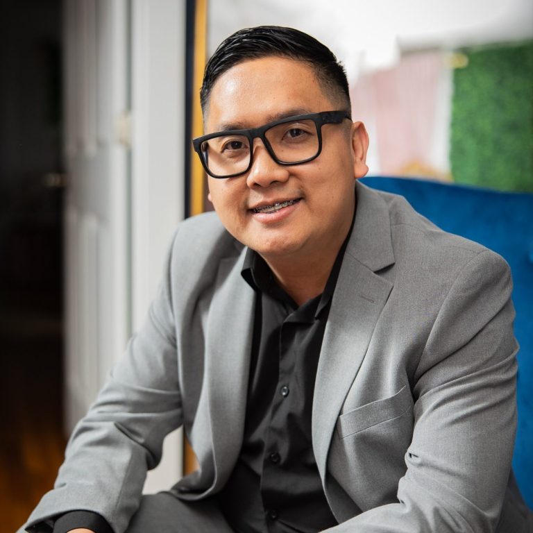 Smiling man in glasses wearing a gray suit and black shirt, sitting on a blue chair.